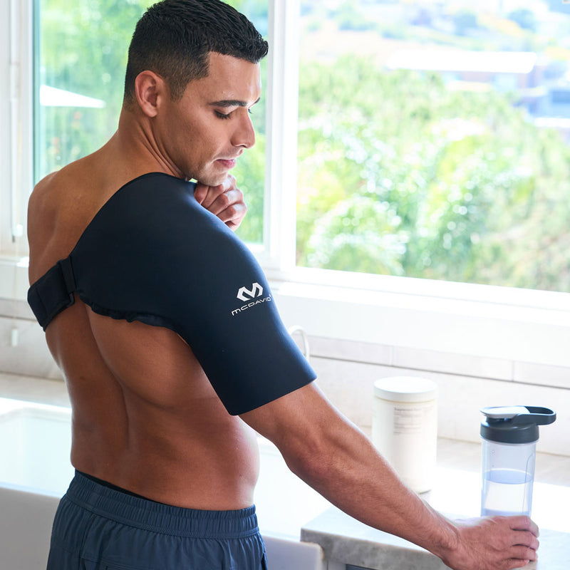Man standing at a kitchen counter wearing the black McDavid Flex Ice Shoulder Sleeve, showing its contoured shoulder coverage and adjustable strap secured across his upper back.