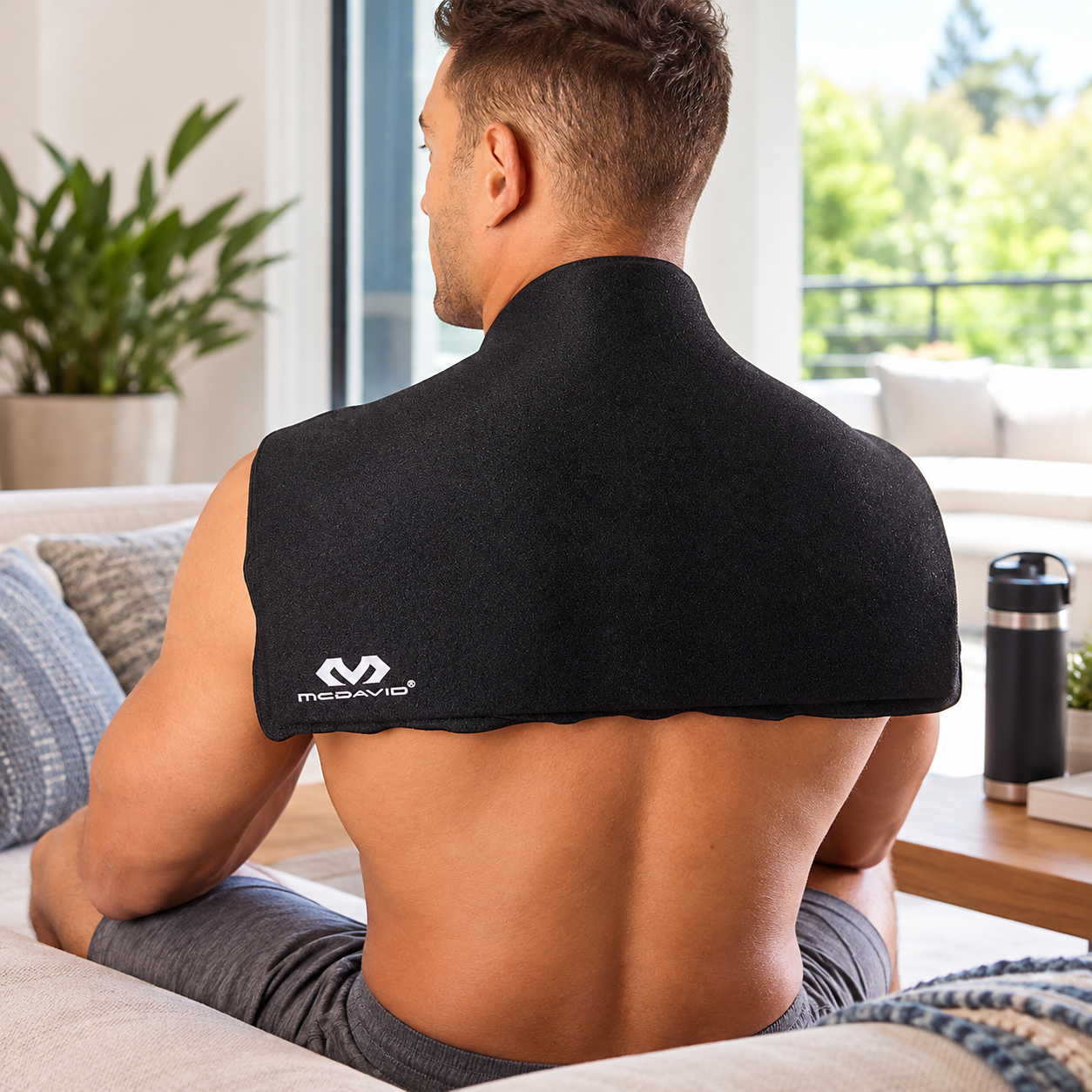 Rear view of a seated man wearing a black McDavid Flex Ice Therapy Blanket draped across his neck, shoulders, and upper back while relaxing on a couch.