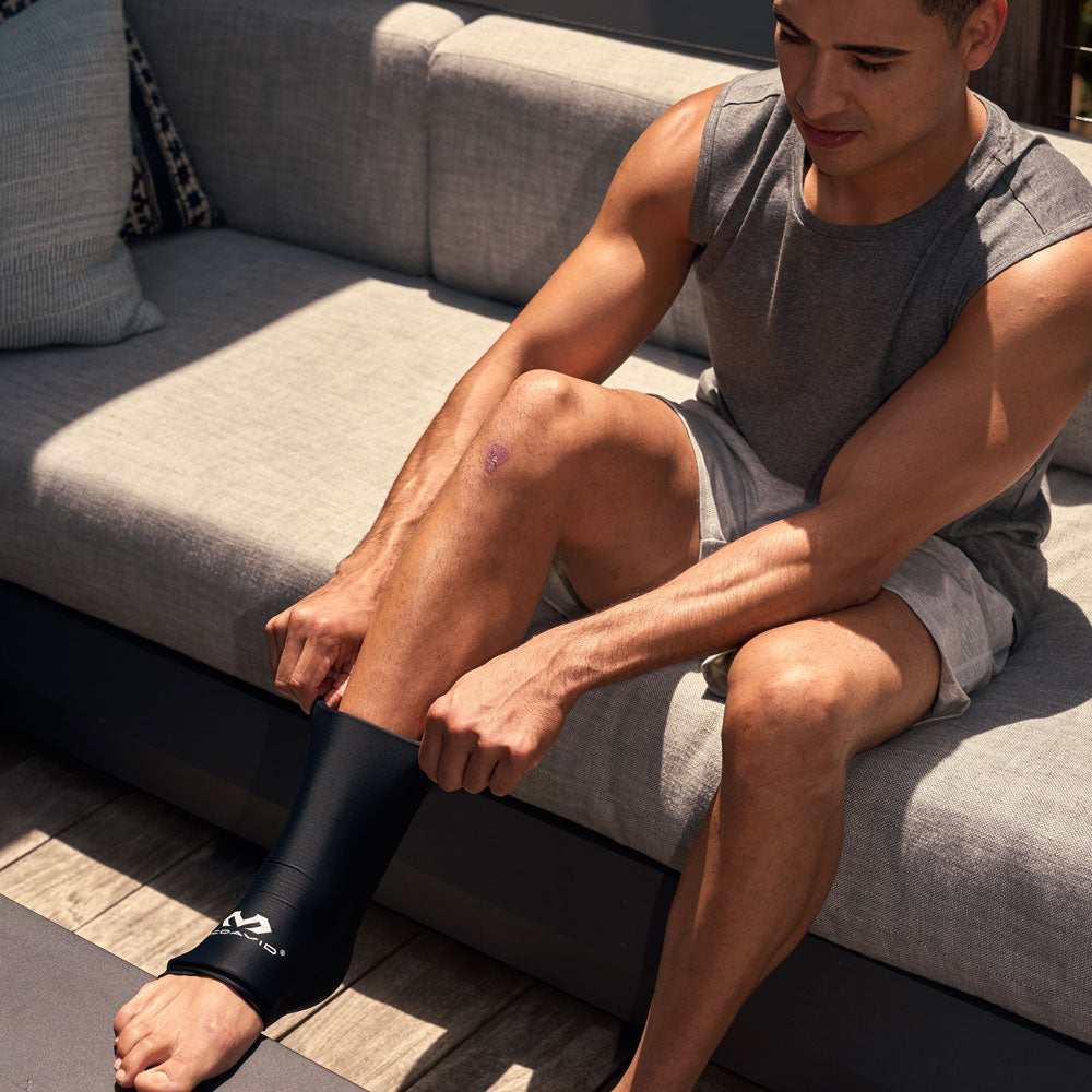 Man sitting on an outdoor couch pulling the black McDavid Flex Ice Ankle Compression Sleeve over his lower leg and ankle for targeted hot or cold therapy.
