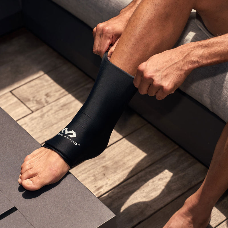 Close-up of a man adjusting the black McDavid Flex Ice Ankle Compression Sleeve, highlighting its flexible material, open-toe design, and coverage around the ankle.