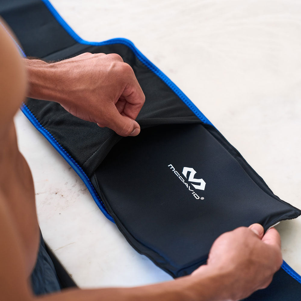 Close-up of a man inserting a black McDavid Flex Ice Therapy Pack into the blue-trimmed back compression sleeve on a countertop
