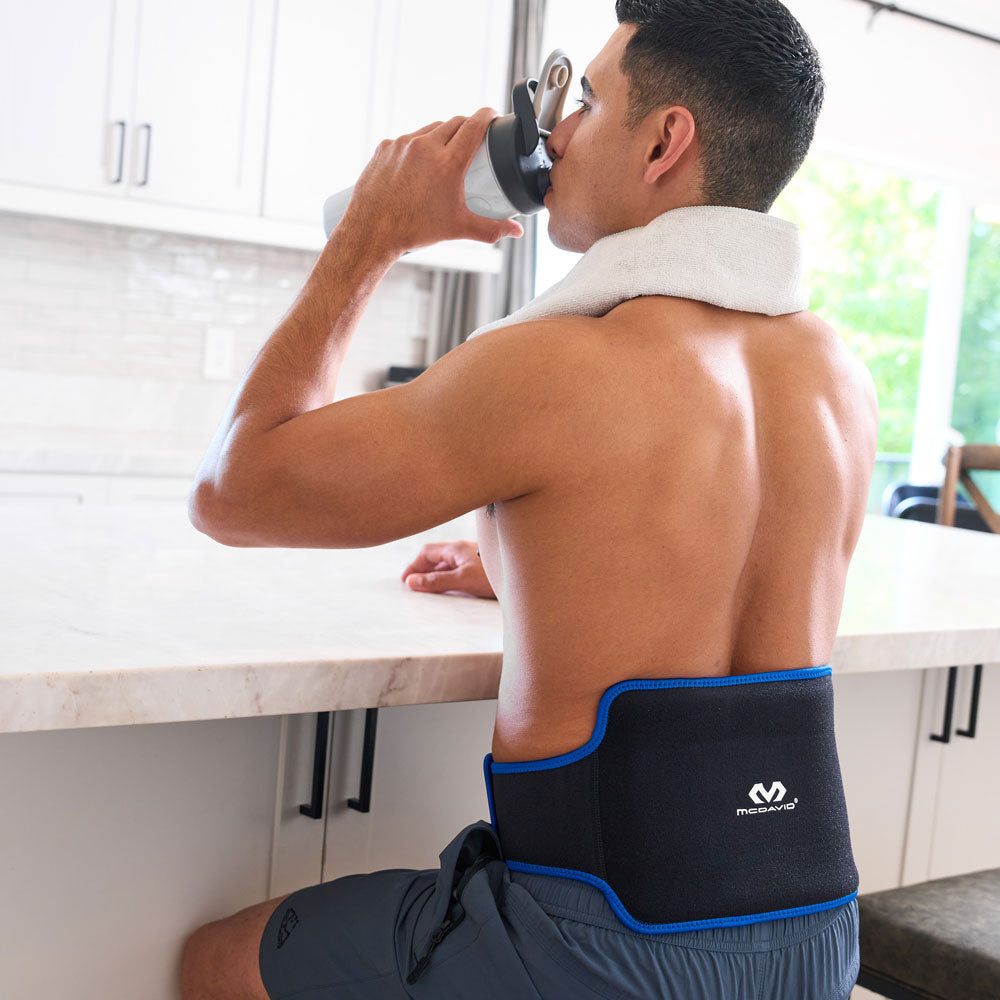Man sitting at a kitchen counter drinking from a water bottle while wearing the black McDavid Flex Ice Back Compression Sleeve with blue trim around his lower back.