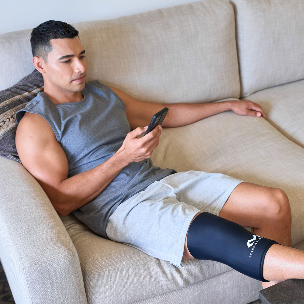 Man reclining on a couch while using his phone and wearing the black McDavid Flex Ice Knee Sleeve for targeted knee therapy.