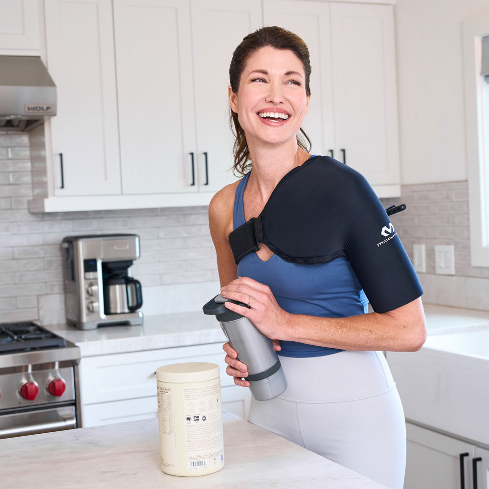 Woman standing in a kitchen holding a shaker bottle while wearing the black McDavid Flex Ice Shoulder Sleeve with an adjustable strap across her upper arm and chest.