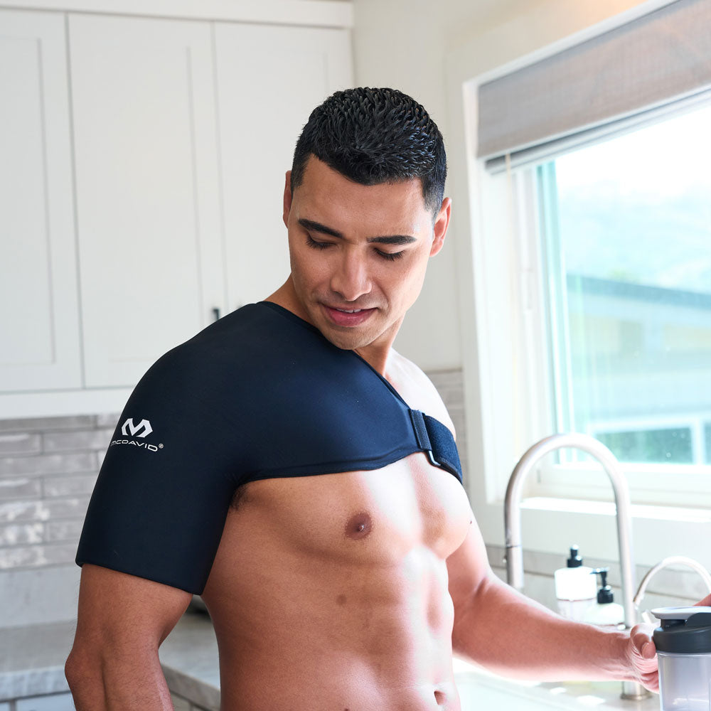 Man standing in a kitchen looking down at the black McDavid Flex Ice Shoulder Sleeve, showing its contoured shoulder and upper arm coverage with an adjustable strap across the chest.