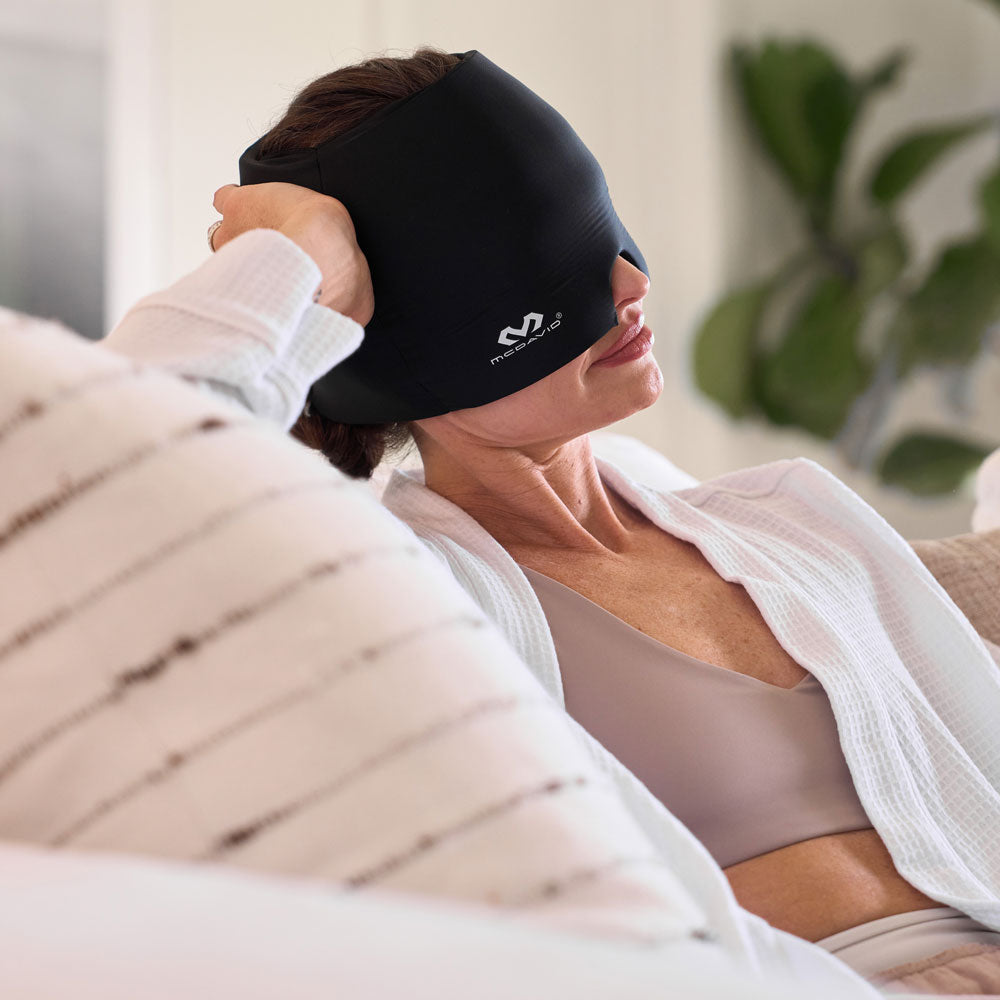 Woman reclining on a couch while wearing the black McDavid Flex Ice Therapy Migraine Cap over her forehead and eyes for soothing hot or cold therapy.