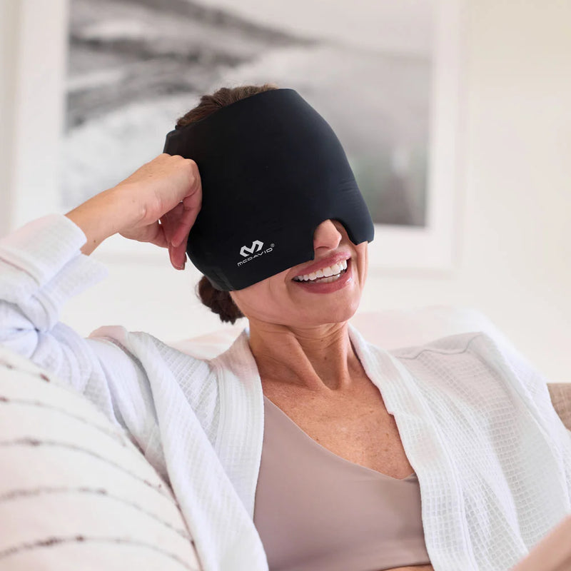 Woman relaxing on a couch in a white waffle-knit robe, smiling while wearing the black McDavid Migraine Cap pulled down over her eyes and forehead.