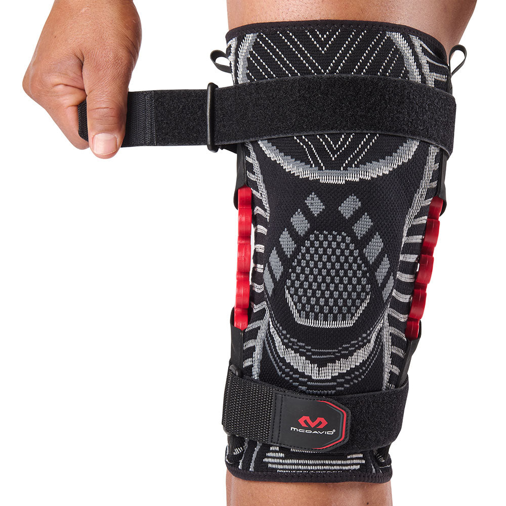 McDavid NRG Lightweight Knit Knee Sleeve with Spring Hinge worn on a person's knee, with the upper adjustable strap being pulled outward to demonstrate the hook-and-loop fastening system. Shows the knit patella support area and bilateral red spring hinges along the sides.