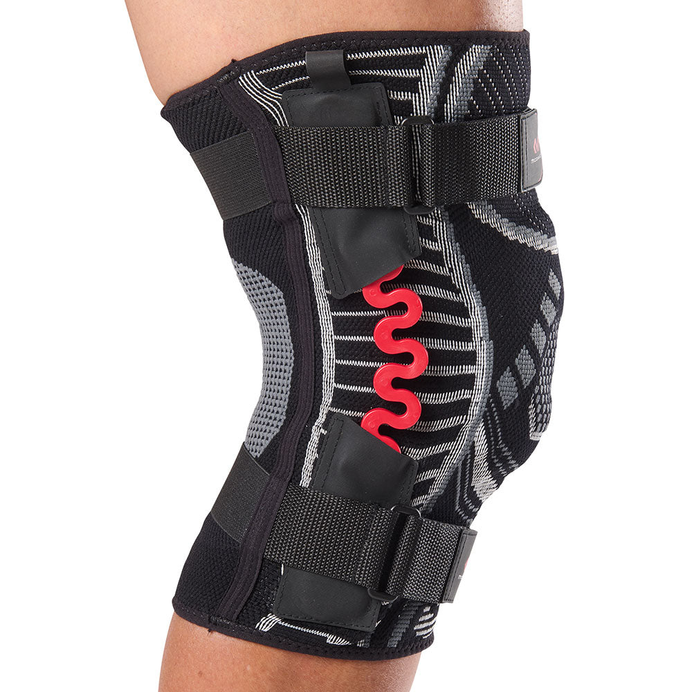 Side view of the McDavid NRG Lightweight Knit Knee Sleeve with Spring Hinge worn on a person's knee, showing the black and grey knit construction, red NRG spring hinge stabilizer along the side of the joint, and upper and lower adjustable compression straps.