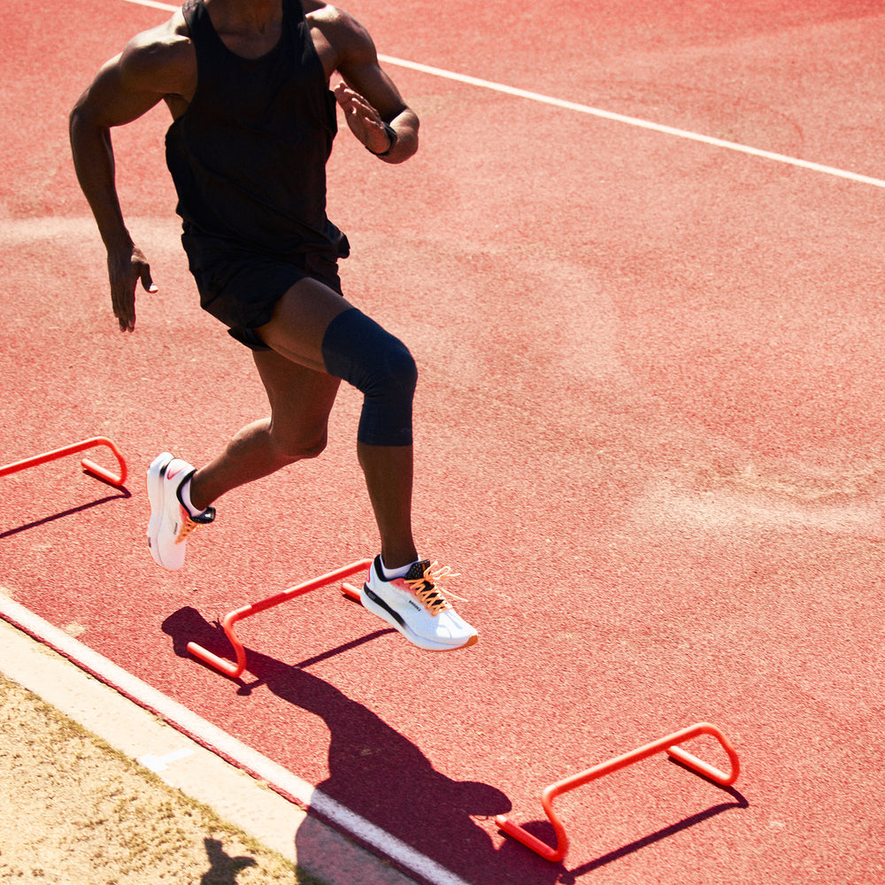 Man sprinting through an agility hurdle drill on a track wearing a navy Infused Buttersoft knee sleeve