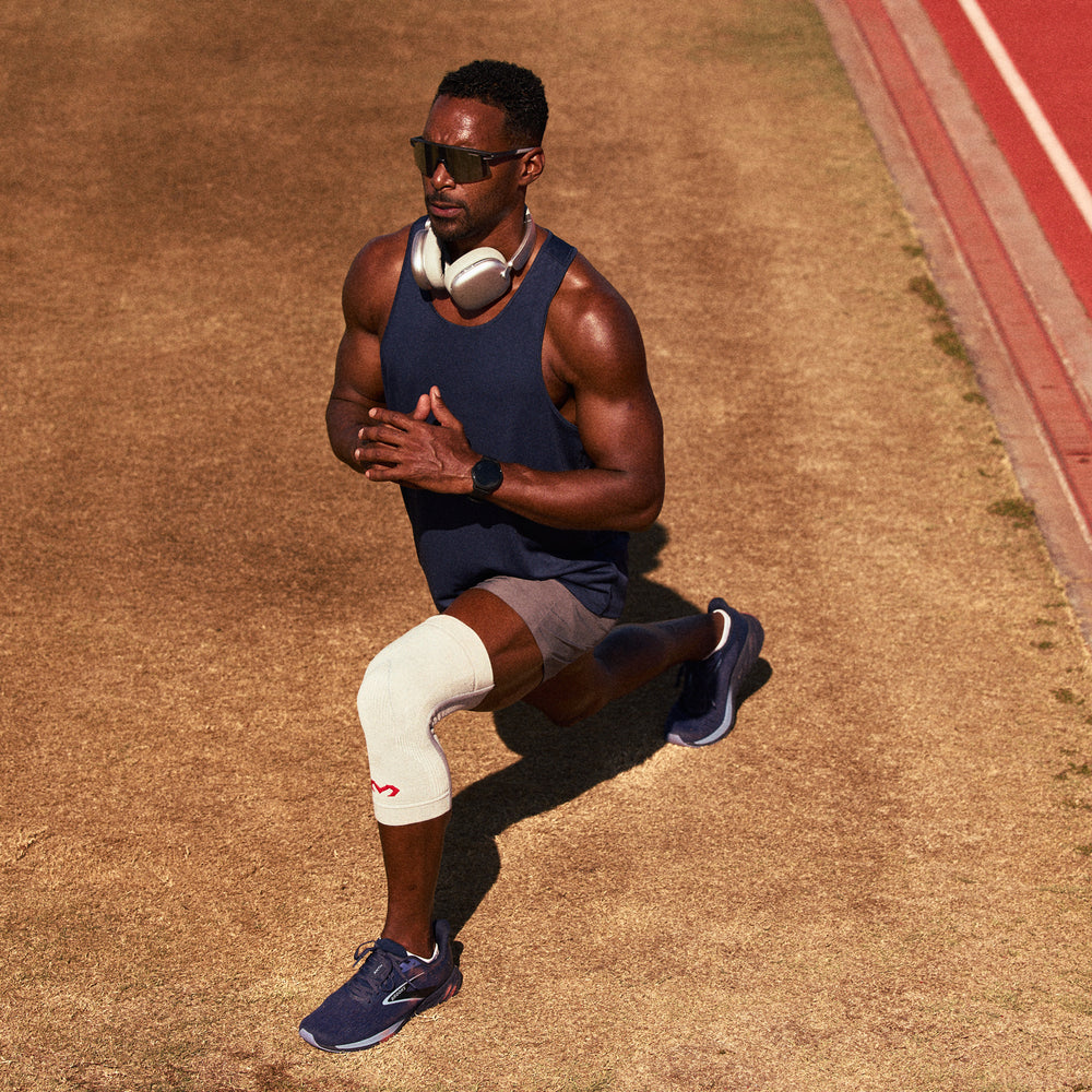 Man jogging on a track wearing a heather gray Infused Knit knee sleeve, sunglasses, and headphones around his neck