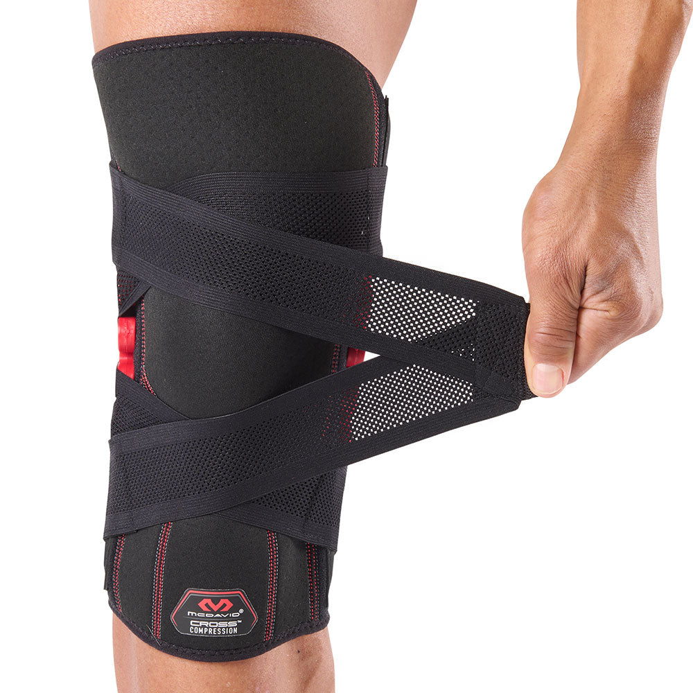 McDavid NRG Cross Compression Knee Sleeve with Spring Hinge worn on a person's right knee, with one of the elastic cross-compression straps being pulled outward to demonstrate the adjustable wrap-around tightening system.