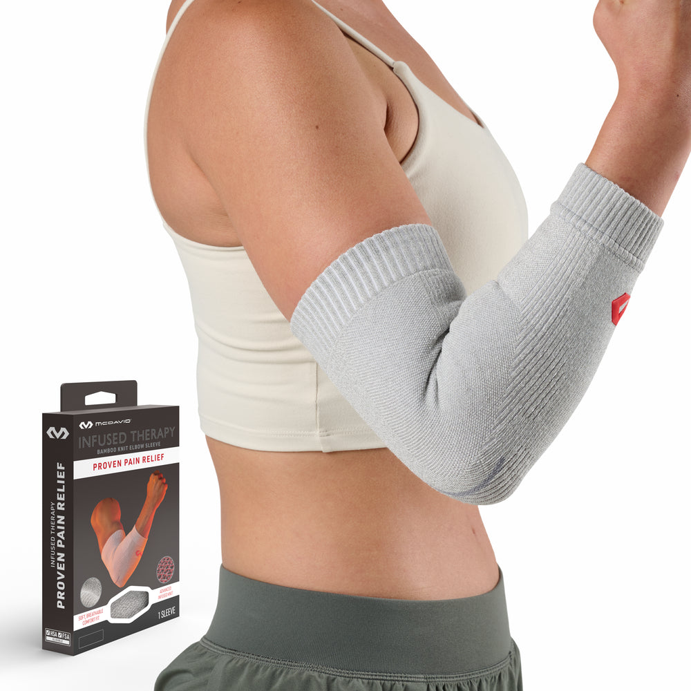 Woman modeling a gray McDavid Infused Therapy bamboo knit elbow sleeve on her bent right arm, standing beside the product packaging labeled 'Infused Therapy Bamboo Knit Elbow Sleeve — Proven Pain Relief.'