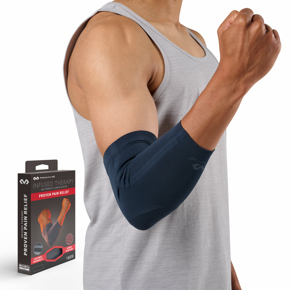 Man wearing a gray tank top with a navy McDavid Infused Therapy buttersoft arm sleeve on his forearm and elbow, fist raised, standing beside the product packaging labeled 'Infused Therapy Buttersoft Arm Sleeve — Proven Pain Relief.'